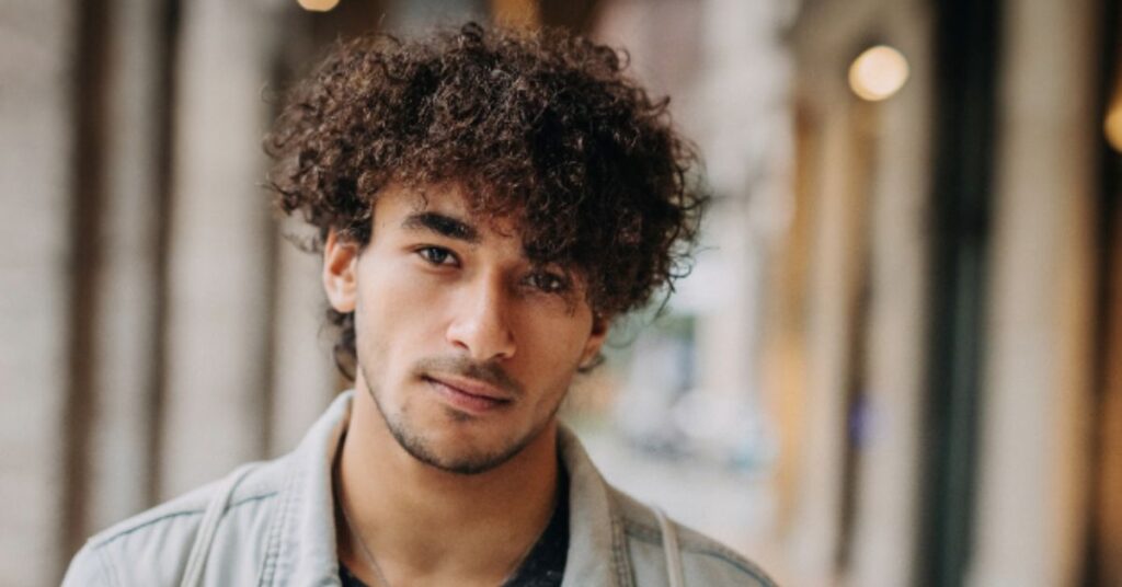 Men’s coily afro shag featuring defined 4C curls, extreme layered volume on top, and tapered sides for a modern round silhouette.