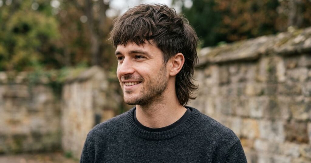Smiling man with a choppy layered wolf cut, a short fringe and longer layers falling over the ears, in a grey knit sweater